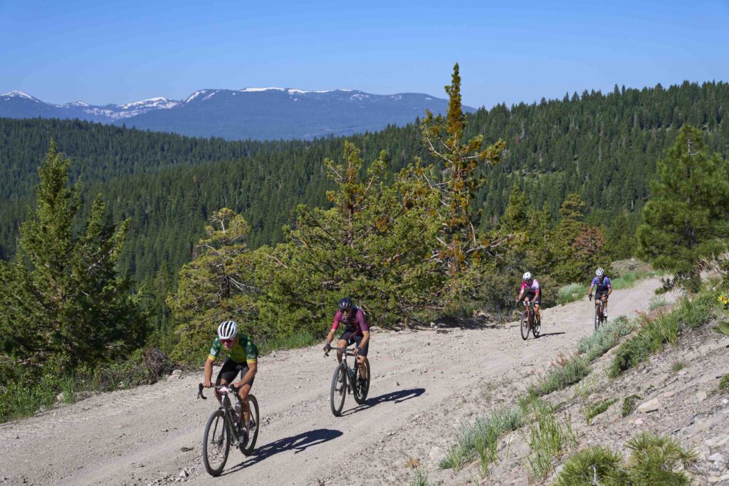 Four cyclists pedaling up a rocky gravel road with the snow covered Sierra Nevada in the distance on a sunny day